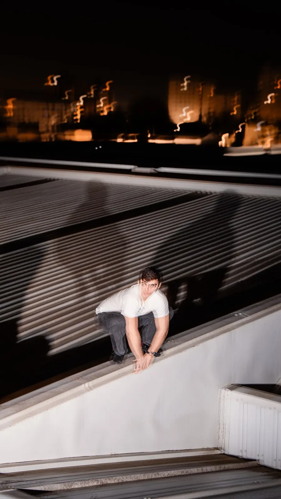 Long-exposure night photo of a Sadhäna band member on a rooftop ledge with city lights blurred behind